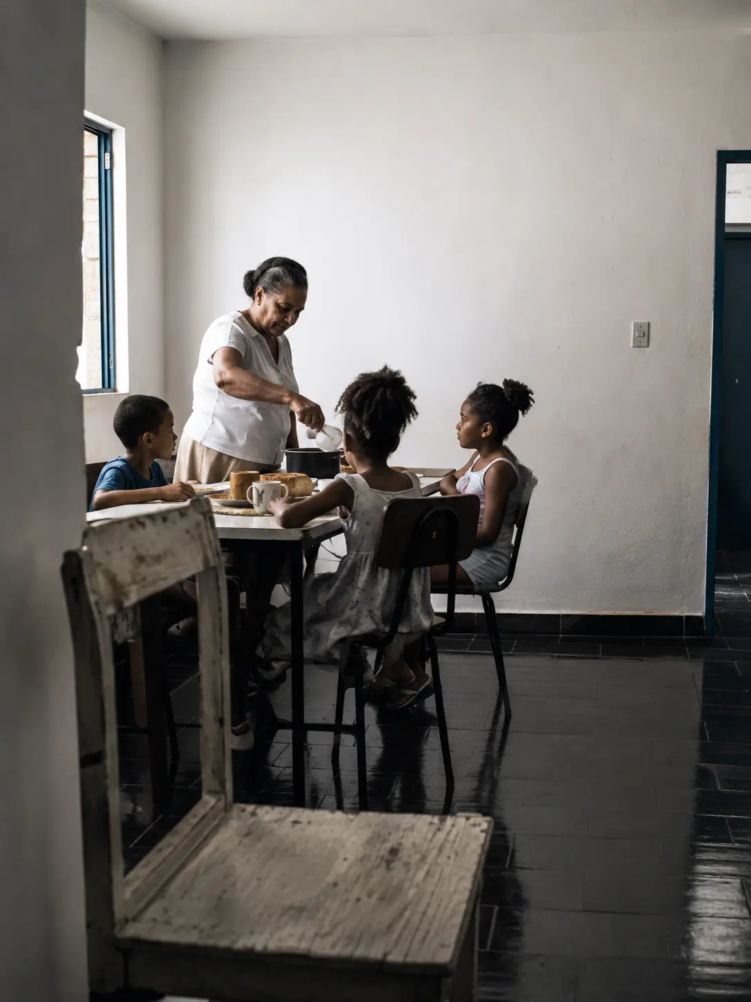 Avó servindo refeição para três crianças em mesa simples.