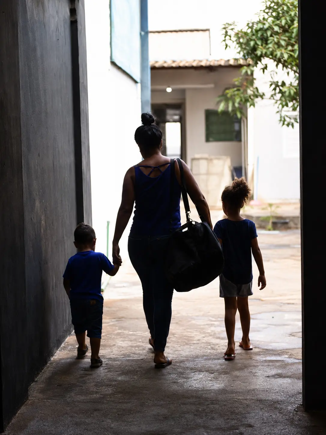 Mãe e duas crianças caminhando por corredor externo ensolarado.