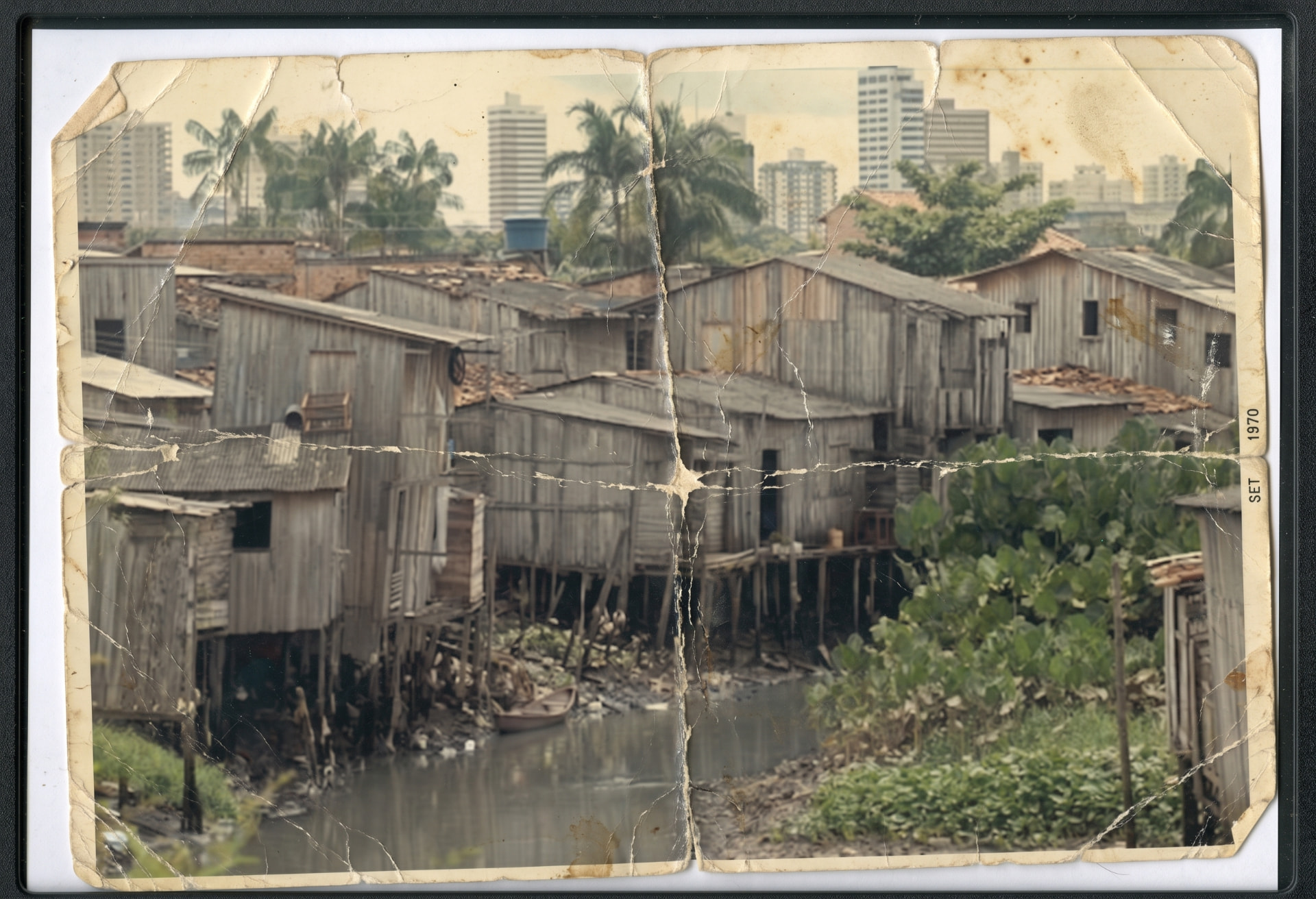 Fotografia antiga e marcada pelo tempo de casas simples de madeira sobre palafitas, com edifícios ao fundo.