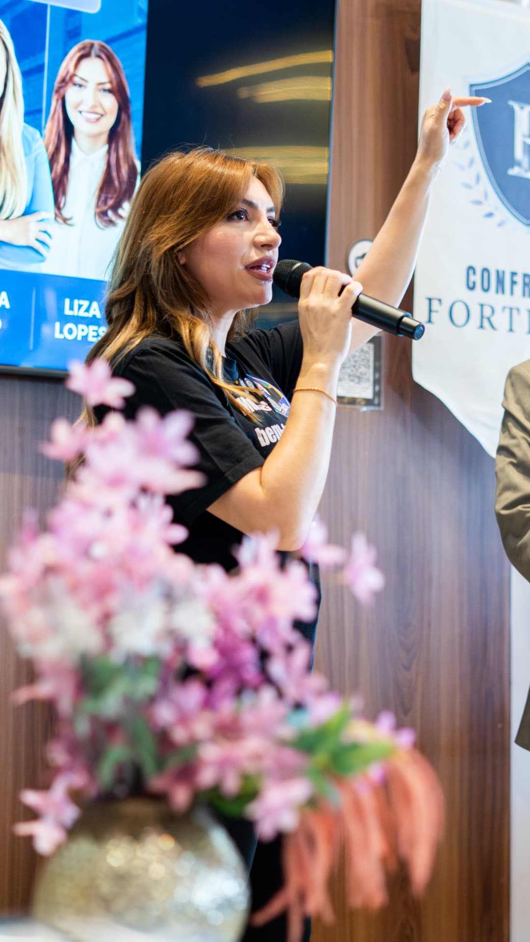 Liza Lopes fala ao microfone durante evento, apontando para um painel institucional.